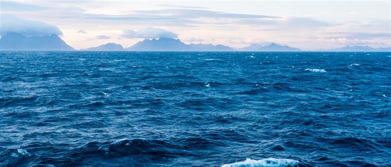 View of a calm blue ocean with gentle waves and distant mountains under a cloudy sky, symbolizing sustainability and innovation in Norway’s aquaculture industry
