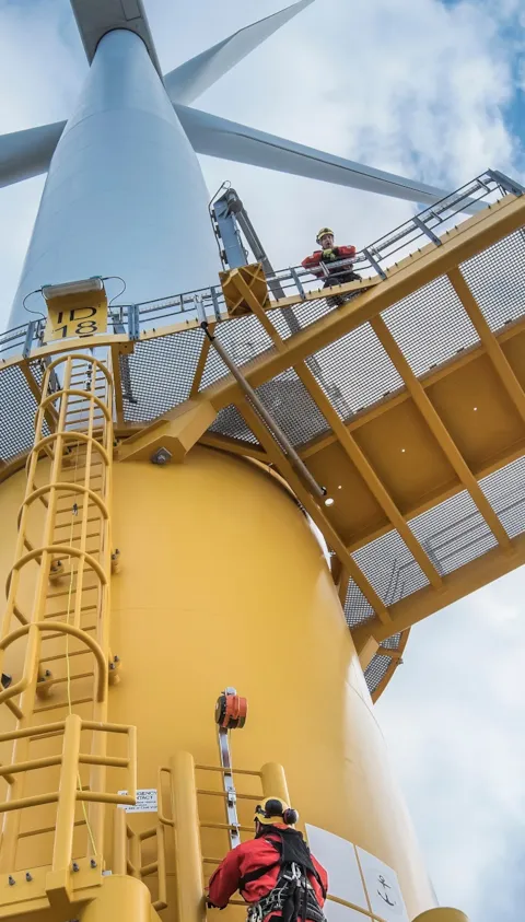 Engineers climbing wind turbine from boat at offshore windfarm, low angle view